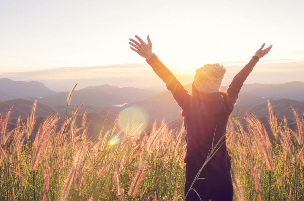 Lady standing in a field of long grass with the sun setting in front of her and her arms spread out in a star shape. SHe is over looking the countryside. 