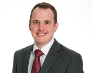 Headshot image of a man wearing a grey suit, white shirt and deep red tie. He is looking at the camera and smiling. The background is white.